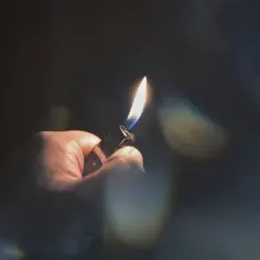 Dłoń trzyma zapaloną zapalniczkę w ciemności. Płomień rzuca ciepłe światło, tworząc refleksy.