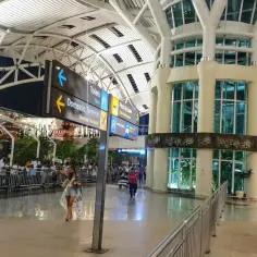 Wnętrze lotniska na Bali. Ludzie z bagażami idą w kierunku znaków: Toalety, Terminal Domowy, Strefa Odbioru.