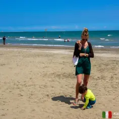 Kobieta i dziecko na piaszczystej plaży w słoneczny dzień, w tle morze i inni plażowicze.