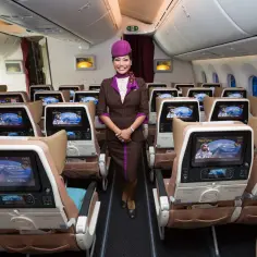 Uśmiechnięta stewardessa Etihad Airways w brązowym mundurku stoi w rzędzie foteli. Ekraniki pokazują astronautę. Dobre etihad airways opinie.