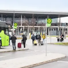 Ludzie z bagażami wsiadają do zielonego autobusu przed nowoczesnym budynkiem, przypominającym jedno z lotnisk w Berlinie.