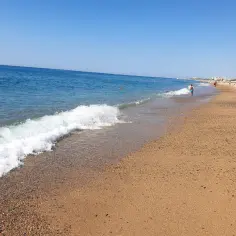 Słoneczny dzień na plaży w Malgrat de Mar. Fale delikatnie obmywają piaszczysty brzeg, a w oddali widać ludzi kąpiących się w morzu.