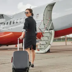 Uśmiechnięta stewardessa z walizką przy samolocie. Czas na boarding, czyli wejście na pokład.