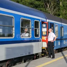 Konduktor w białej koszuli stoi przy drzwiach niebieskiego pociągu InterCity na peronie.