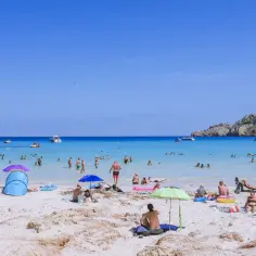 Słoneczna plaża Cala Agulla tętni życiem. Ludzie kąpią się w krystalicznie czystej wodzie, opalają na piasku pod parasolami i relaksują na leżakach.