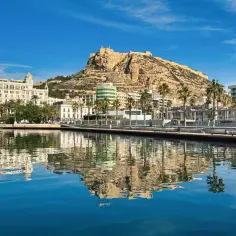 Piękna pogoda w Alicante w marcu. Błękitne niebo, spokojna woda odbijająca port, białe jachty i majestatyczny zamek Santa Barbara na wzgórzu.