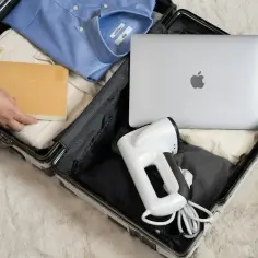 Pakowanie bagażu rejestrowanego: laptop, parownica do ubrań i książka obok niebieskiej koszuli. Elektronika w bagażu rejestrowanym to wygoda w podróży.