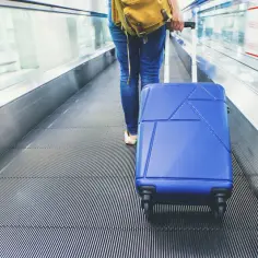 Osoba z niebieską walizką na ruchomych schodach lotniska. Zastanawiasz się, ile kosztuje odprawa na lotnisku?