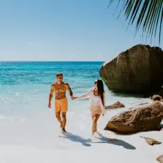 Para, trzymając się za ręce, spaceruje po plaży na Seszelach. Woda jest turkusowa, a na brzegu leżą kamienie.