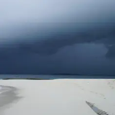 Malediwy pora deszczowa. Ciemne chmury nad turkusową wodą i białą plażą. Słomiany parasol czeka na turystów.
