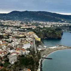 Widok na malownicze miasteczko nad morzem, z klifami schodzącymi do wody. W oddali widać góry. To idealne miejsce na wypoczynek, jak na neapol plaża.