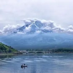 Majestatyczny Fudżi, otoczony chmurami, góruje nad jeziorem. W oddali most, a na wodzie łódka z wędkarzami. Zastanawiasz się, ile się leci do Japonii?