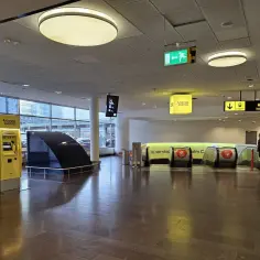 Żółte automaty biletowe Arlanda Express na lotnisku w Sztokholmie. W tle ruchome schody i znaki kierujące.
