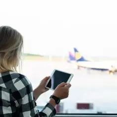 Kobieta z tabletem przy oknie lotniska, sprawdza, na czym polega odprawa online, zanim wsiądzie do samolotu.