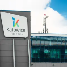 Logo Katowice Airport na szarym budynku, obok przeszklona część i maszt telekomunikacyjny.