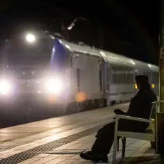 Osoba siedzi na ławce na peronie nocą, zbliża się pociąg z zapalonymi światłami.