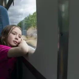 Dziewczynka wpatruje się przez okno pociągu, zastanawiając się, od ilu lat można jeździć samemu pociągiem.
