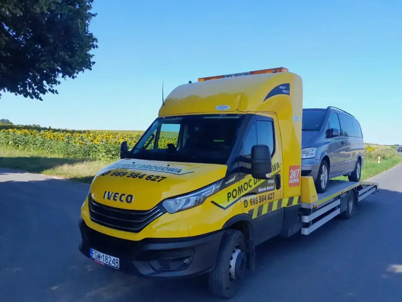 Żółta laweta z autem na pokładzie, gotowa do assistance krótkoterminowe 1500 km. Tło to pole słoneczników.