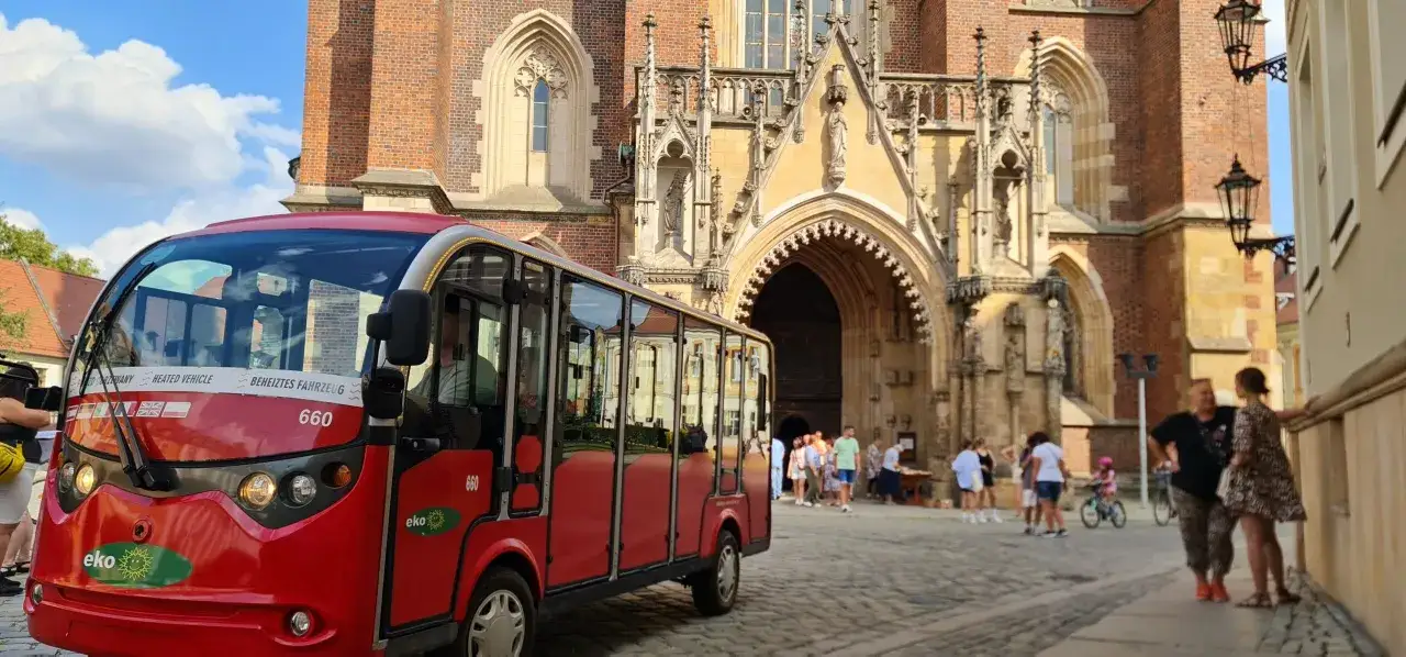 Czerwony, elektryczny pojazd turystyczny czeka na pasażerów przed zabytkową katedrą, idealny na city break z Wrocławia.