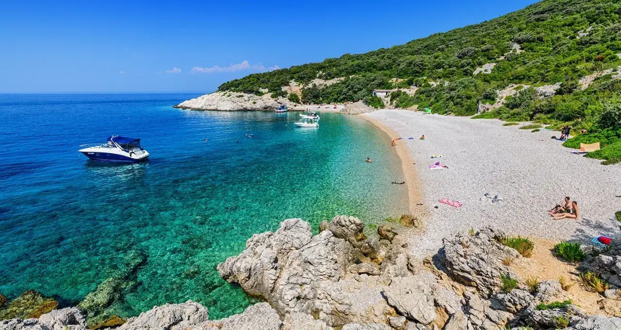 Idealne miejsce na wycieczki do Chorwacji: kamienista plaża, turkusowa woda, łódź i zielone wzgórza.