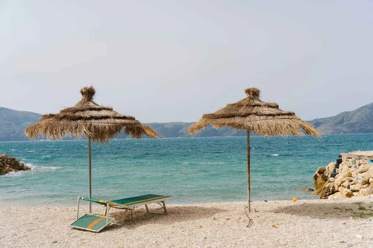 Dwa parasole plażowe i leżaki nad turkusowym morzem. Widok gór w tle. Czy w Albanii jest bezpiecznie? Tak, to spokojne miejsce na wypoczynek.