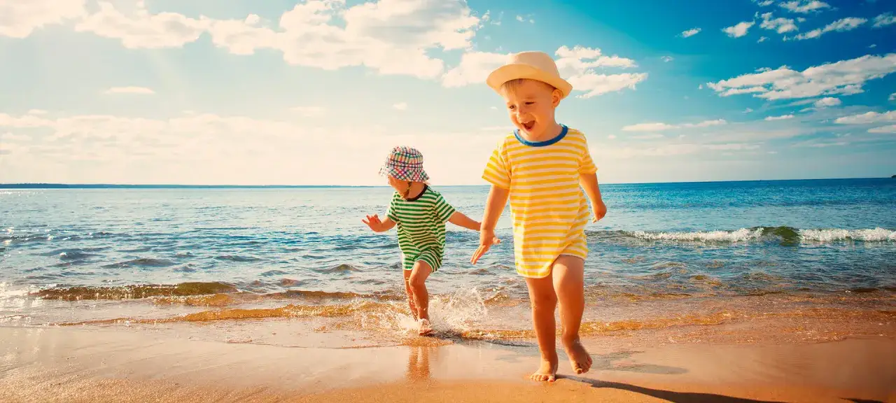 Dzieci biegają po plaży w Malgrat de Mar, chlapiąc w wodzie. Słońce świeci, a niebo jest błękitne z białymi chmurami.