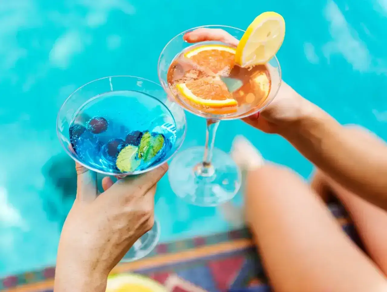 Dwie ręce trzymają kieliszki z kolorowymi drinkami przy basenie. Idealny all inclusive soft na wakacjach.