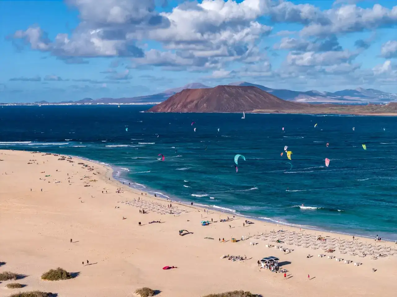 Maj na Fuerteventurze to idealna pogoda na kitesurfing. Widać plażowiczów i kolorowe latawce na turkusowym morzu.